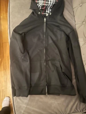 Svart hoodie  - Säljer en svart hoodie med dragkedja och fickor framtill. Huvan har ett rutigt innerfoder i svart, vitt och rött som ger en snygg kontrast. Klassisk passform och ribbade muddar vid ärmar och nederkant. Perfekt för en chill och stilren look.