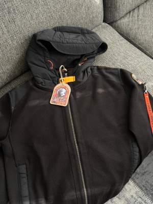 Parajumpers jacka - Helt nyrr Snygg svart vindjacka från Parajumpers med huva, dragkedja och orange detaljer. Jackan har en patch på ärmen och praktiska fickor. Perfekt för dig som vill ha en sportig och stilren look. Storlek S