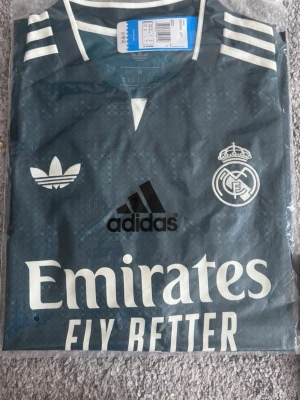 Grön Real Madrid tröja Adidas - Snygg grön Real Madrid matchtröja från Adidas med vita detaljer och klubbmärke på bröstet. Tröjan har klassiska Adidas-ränder på axlarna och trycket 'Emirates Fly Better' på framsidan. Tillverkad i lätt och ventilerande material.