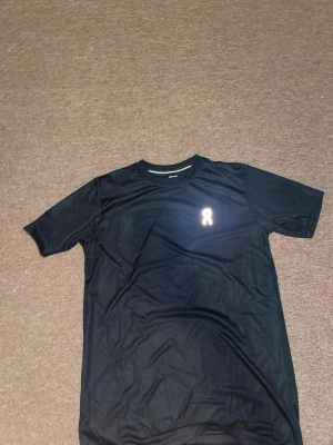 Svart sportig t-shirt med reflexdetalj - Säljer en svart sport-tshirt med rund hals och korta ärmar. Materialet är lätt och andas, perfekt för träning. På bröstet finns en reflexdetalj som ger extra synlighet. Passar dig som gillar en stilren och funktionell look på gymmet.