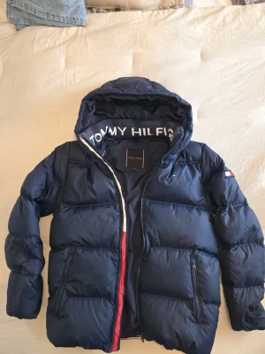 Tommy Hilfiger Puffer Jacket - This is a navy blue puffer jacket from Tommy Hilfiger. It features a hood with 'TOMMY HILFIGER' branding on the inside, a front zipper, and zippered side pockets. The jacket also has the Tommy Hilfiger flag logo on the left sleeve and a branded label inside the collar area. The size is 16Y, which is typically for youth/teens.