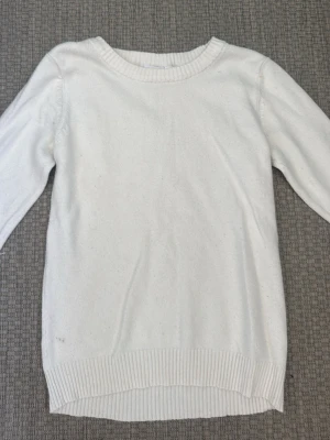 Vit stickad tröja med rund hals - Klassisk vit stickad tröja med rund halsringning och långa ärmar. Tröjan har ribbstickade muddar vid ärmslut och nederkant, vilket ger en snygg och enkel look. Perfekt att matcha med jeans eller kjol för en clean stil.