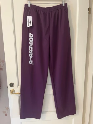Lila retro byxor från Bagheera strl XL - Snygga lila retro mjukis/tränings byxor från Bagheera med vitt tryck längs ena benet. Byxorna har resår i midjan, en bakficka med dragkedja och två framfickor med dragkedja. Ny med prislapp, aldrig använd. Storlek XL.