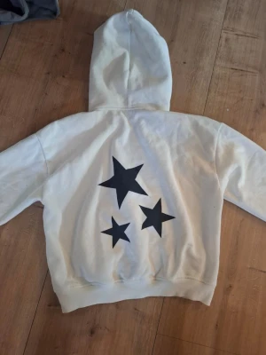 Vit hoodie med svarta stjärnor XS - Säljer en vit hoodie i storlek XS med tre stora svarta stjärnor tryckta på ryggen. Hoodien har klassisk huva och ribbade muddar. Perfekt för dig som gillar enkel men cool design. Materialet är mjukt och skönt, troligen bomull eller bomullsblandning.
