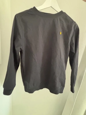 Svart sweatshirt med gul logga - En svart sweatshirt från lyle & scoot med rund hals och lång ärm. Tröjan har en liten gul broderad logga på bröstet och ribbade muddar vid ärmslut och nederkant. Tillverkad i mjukt bomullsmaterial för en skön känsla. I storlek 12-13 år 