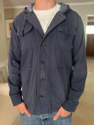 Marinblå hooded overshirt från Vans - Snygg marinblå overshirt med huva och knappar framtill. Jackan har stora fickor både på bröstet och nedtill samt dragsko i huvan. Tillverkad i bomull och passar perfekt till vardagslooken.