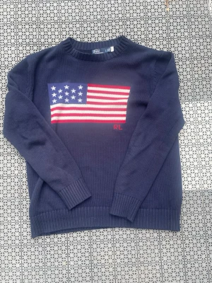 Marinblå stickad tröja Polo Ralph Lauren - Stickad marinblå tröja från Polo Ralph Lauren med amerikansk flagga i rött, vitt och blått på bröstet samt RL-broderi. Tröjan har rund halsringning och ribbade muddar vid ärmslut och nederkant. Tillverkad i bomull för en mjuk känsla. Passar dig som är runt 180
