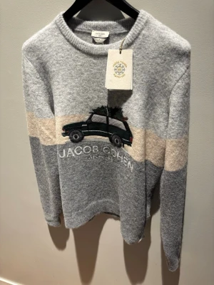 Grå stickad tröja Jacob Cohën - FJERNER OM 6 TIMAR. Helt ubrukt og trenger pengene                                   Ai: Stickad tröja i grått med beige band och broderad bil med gran på taket framtill. Mjuk ullmix, rund hals och ribbade muddar. Snygg detalj med texten 'Jacob Cohën Après Ski' under motivet. Perfekt för dig som gillar unik design.