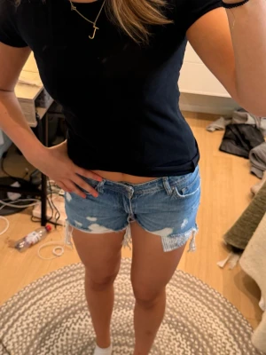 Blå slitna jeansshorts med fransar - Säljer ett par blå jeansshorts med slitna detaljer och fransar längs bensluten. Shortsen har klassisk femficksmodell, knapp och dragkedja framtill samt råa kanter för en avslappnad look. Perfekta för varma dagar och enkel att matcha.