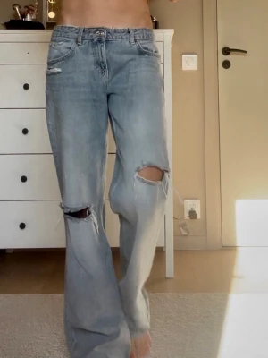 Ljusblå slitna zara jeans low waist straight - Säljer ett par ljusblå straight low waist jeans från zara med stora slitningar vid knäna och avslappnad passform. Jeansen har klassisk femficksdesign och är tillverkade i denim. Perfekta för en trendig och avslappnad look.