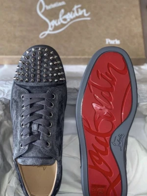 Gråa Louboutin med spikar - Säljer ett par gråa sneakers från Christian Louboutin i mocka med ikoniska röda sulor och silverfärgade nitar på tån. Skorna har snörning och låg profil, samt detaljer i metall. Perfekta för dig som vill sticka ut med en edgy look.