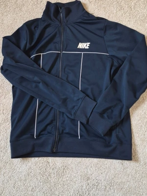 Mörkblå zip-jacka från Nike - Snygg mörkblå träningsjacka från Nike med vit logga på bröstet och vita detaljer längs sömmarna. Jackan har hel dragkedja, hög krage och är tillverkad i ett mjukt, stretchigt material som passar perfekt till träning eller chill.