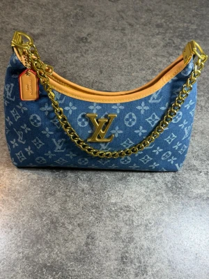 Blå Louis Vuitton denim hand och axel väska  - Snygg handväska i blå denim med klassiskt Louis Vuitton-mönster och guldfärgad kedja. Väskan har detaljer i ljust läder och ett stort LV-emblem framtill. Perfekt accessoar för dig som vill sticka ut med en trendig och exklusiv look.