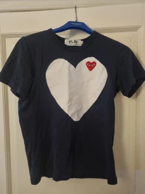Blå Comme des Garçons Play t-shirt - Mörkblå t-shirt från Comme des Garçons Play med stort vitt hjärta och röd hjärtdetalj med ögon på bröstet. Klassisk rund hals och korta ärmar. Perfekt för dig som gillar streetwear och vill sticka ut med en ikonisk design. OBS! Den sitter som S/Xs.