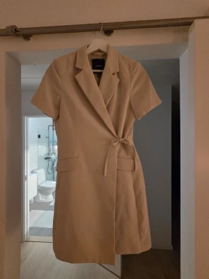 Beige omlottklänning med knytband - Stilren beige kortklänning med omlott-design och knytband i midjan. Klänningen har korta ärmar, klassisk krage och två fickor framtill. Perfekt för en chic och enkel look.
