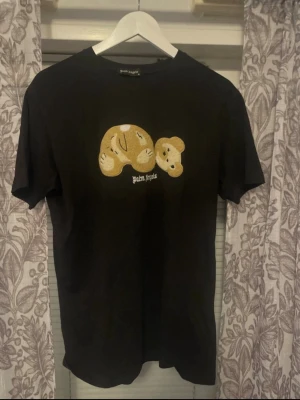 Svart Palm Angels t-shirt med björn - Svart t-shirt från Palm Angels med ikoniskt björnmotiv på bröstet och vit logga under. Klassisk rund hals och korta ärmar. Perfekt för dig som vill ha en streetwear-look med edge. XL men passar till L