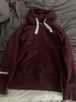 Vinröd hoodie med dragkedja Russedress - Säljer en vinröd hoodie från Russedress med dragkedja framtill, vita snören i huvan och fickor på sidorna. Mjuk och skön bomullsmix, perfekt för chill dagar. Logga på ärmen för extra stilpoäng.