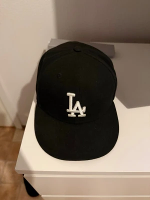 Svart LA keps från New Era - Säljer en svart keps från New Era med LA-logga broderad i vitt framtill. Klassisk 59FIFTY-modell med rak skärm och strukturerad passform. Kepsen är tillverkad i bomull och polyester, perfekt för en streetwear-look.