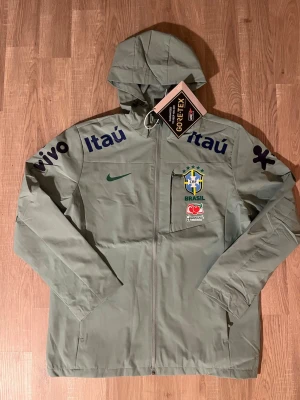 Grön Brasilien vindjacka - Storleken på jackan är XL MEN SITTER SOM STORLEK S!!! Snygg grön vindjacka med Brasilien-logga och sponsormärken som Itaú och Vivo. Jackan har huva, hel dragkedja och är tillverkad i GORE-TEX-material som står emot regn och vind. Perfekt för dig som gillar sportig stil och vill visa stöd för Brasilien. HELT NY!