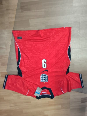 England fotbollströja röd Kane #9 - Snygg röd England fotbollströja med svarta och vita detaljer, nummer 9 och namnet Kane på ryggen. Tröjan har korta ärmar, broderad landslagslogga och Nike-logga på framsidan. Perfekt för dig som älskar fotboll och vill stötta England.