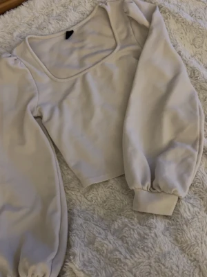Beige croppad tröja med puffärm - Trendig beige croppad tröja med bred fyrkantig ringning och långa puffärmar. Tröjan har ribbade muddar vid ärmslut och är gjord i ett mjukt, stretchigt material. Perfekt för en stilren och modern look.
