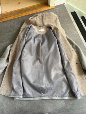 Beige ullkappa från Féraud Paris - Stilren beige ullkappa från Féraud Paris med klassisk rak passform och grått innerfoder. Kappan har huva och dolda knappar framtill. Perfekt för kyliga dagar och ger en tidlös look till din outfit. Den ena knappen har gått av. 