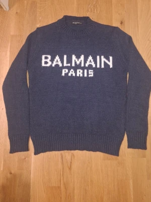 Balmain tröja  - Snygg mörkblå stickad tröja från Balmain med vit text 'BALMAIN PARIS' framtill. Tröjan har rund halsringning, ribbade muddar vid ärmslut och nederkant. Perfekt för dig som gillar exklusiva och trendiga plagg.