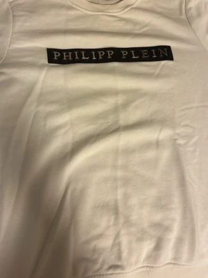 Vit t-shirt med logga - Snygg vit t-shirt  med svart logga framtill. Klassisk rund halsringning och korta ärmar. Tillverkad i mjukt bomullsmaterial som känns skönt mot huden. Perfekt för dig som gillar stilrena och exklusiva plagg.