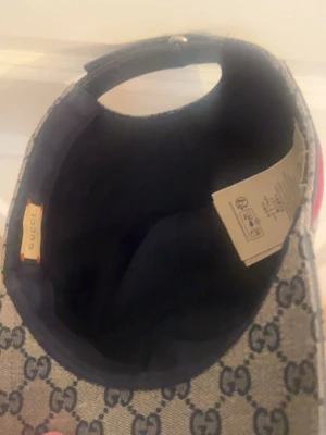 Gucci keps med GG-mönster - Snygg keps från Gucci i beige tyg med klassiskt GG-monogram över hela kepsen. På sidan finns en röd och marinblå rand som ger extra stilpoäng. Kepsen har böjd skärm och justerbar rem baktill. Perfekt för dig som vill sticka ut med en lyxig accessoar.