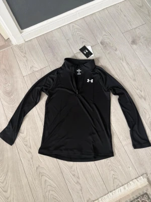 Under armour - Svart tränings-/underställströja i nytt skick. Stilren modell med dragkedja och Under armour logga. Ny med tags kvar