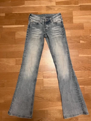 Ljusblå bootcut jeans med slitningar - Snygga ljusblå jeans med bootcut-modell och slitna detaljer framtill. Jeansen har klassisk femficksdesign och en grön patch baktill i midjan. Perfekta för en avslappnad och trendig look.