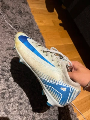 Nike fotbollsskor med blå detaljer - Snygga fotbollsskor från Nike i vitt med blå detaljer och stora Nike-loggor på sidorna. Skorna har snörning och en låg profil för bra bollkontroll. Tillverkade i syntetmaterial som ger en lätt känsla på planen. Perfekta för dig som vill sticka ut på matchen.