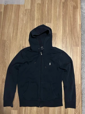 Svart hoodie från Polo Ralph Lauren - Snygg svart hoodie från Polo Ralph Lauren med dragkedja framtill och klassisk vit logga på bröstet. Hoodien har huva med dragsko och en stor magficka. Tillverkad i mjukt bomullsmaterial som är skönt att ha på sig. Perfekt för en chill och stilren look.