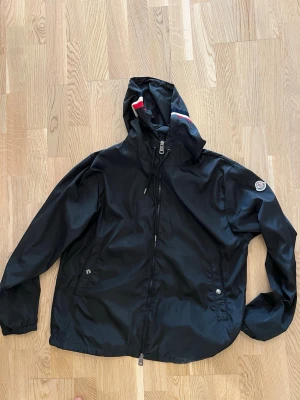 Svart vindjacka med huva Moncler - Snygg svart vindjacka från Moncler med huva och dragkedja framtill. Jackan har två fickor med tryckknappar och Moncler-logga på ärmen. Detaljer i rött och vitt på huvan ger en sportig touch. Tillverkad i lätt och tåligt material, perfekt för blåsiga dagar.