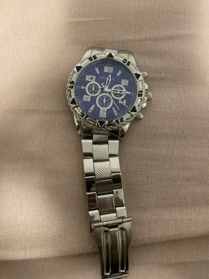 Silverfärgad klocka med blå urtavla - Stilren klocka i silverfärgat metallarmband och blå urtavla med flera små urtavlor och tydliga siffror. Klockan har en sportig look med markerad bezel och robust länkarmband. Perfekt accessoar för dig som gillar en klassisk men modern stil.