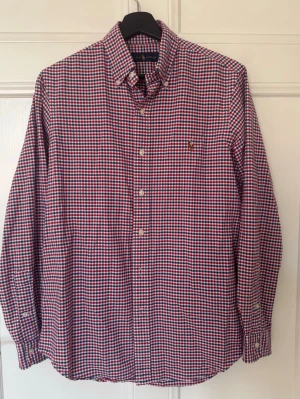 Rutig skjorta från Ralph Lauren - Klassisk rutig skjorta från Ralph Lauren i rött, blått och vitt. Skjortan har knappar framtill, button-down krage och en liten broderad logga på bröstet. Tillverkad i mjuk bomull och har långa ärmar med manschettknappar.