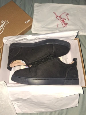 Svarta sneakers från Christian Louboutin - Stilrena svarta sneakers från Christian Louboutin i mocka med matchande svarta skosnören och sula. Låg modell med diskret logga på hälen och klassisk röd detalj på insidan. Perfekta för dig som gillar exklusiv och clean stil.