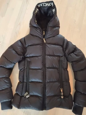 Svart Moncler jacka - Snygg svart dunjacka från Moncler med stor huva och logotyp på huvkanten. Jackan har två stora dragkedjeförsedda fickor framtill och quiltad design. Perfekt för kalla vinterdagar och riktigt varm tack vare dunfyllningen.