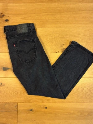 Levi’s 501 W32 L30 - Klassiska Levi’s 501 i tvättad svart denim med en snygg, urblekt patina. Rak passform (straight fit) med normal midjehöjd och knappgylf i äkta Levi’s-stil.  Skick: Fint begagnat skick – naturlig blekning i tvätten ger ett äkta, avslaget uttryck utan större skador, hål eller fläckar. Ficklinjer och söm i gott skick.  Detaljer:  * Modell: 501, rak passform * Färg: Svart/antracit, blekt tvätt * Storlek: W32 L30 * Äkta läderlapp bak samt röd Levi’s-tab på bakfickan * Knappgylf * 100% bomull (denim)