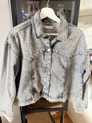Grå jeansjacka med nitar från Zara - Snygg grå jeansjacka från Zara med tvättad look och silverfärgade nitar längs bröstet. Jackan har klassisk krage, knappstängning framtill och raka ärmar. Perfekt för dig som vill ha en trendig och edgy stil.