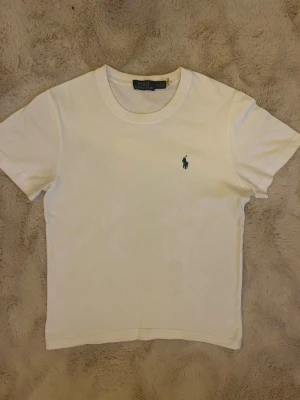 Vit t-shirt från Polo Ralph Lauren - Klassisk vit t-shirt från Polo Ralph Lauren med den ikoniska broderade loggan på bröstet. T-shirten har rund halsringning och korta ärmar. Perfekt basplagg i mjuk bomull som passar till allt. Lite för liten för att vara en S jag tycker mer den sitter som en XS, andvänd fåtal gånger under förra året har en liknande i svart också som jag kan fixa bra pris tillsammans (kolla min andra annons för att se den)