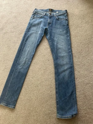 Blå lee jeans - Säljer ett par klassiska blå jeans med slim passform . Jeansen har normal midja, dragkedjegylf och subtila slitningar för en avslappnad look. Tillverkade i denim med snygga kontrastsömmar. Perfekta till en enkel t-shirt eller hoodie.