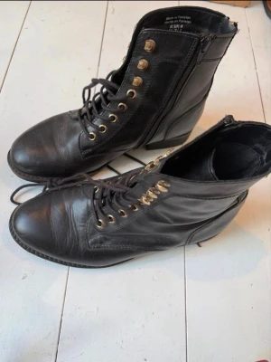 Svarta boots med snörning och dragkedja - Säljer ett par svarta boots i skinn med snörning framtill och guldfärgade detaljer. Skorna har dragkedja både på sidan och baktill för enkel på- och avtagning. Klassisk rund tå och låg klack gör dem perfekta till många olika stilar. Stl 38 men en liten 38. 