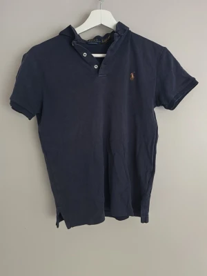 Marinblå pikétröja från Polo Ralph Lauren - Klassisk marinblå pikétröja från Polo Ralph Lauren med korta ärmar och den ikoniska broderade loggan på bröstet. Tröjan har krage och knappar vid halsen. Perfekt till jeans eller shorts för en avslappnad men snygg look. Väldigt skön!