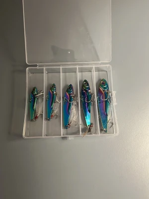 Fiskedrag set i regnbågsfärger - Säljer ett set med fem metallfärgade fiskedrag i olika storlekar, levereras i en genomskinlig plastask med fem fack. Drag i skimrande regnbågsfärger, varje drag har ögon och två krokar. Perfekt för fiskeentusiasten som vill ha snygga och effektiva beten.