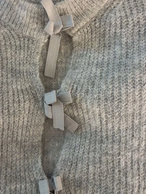 Ljusgrå stickad tröja med rosetter - Superfin ljusgrå stickad tröja med unika rosettdetaljer framtill. Tröjan har rund halsringning och långa ärmar. Materialet ser mjukt och fluffigt ut, perfekt för kyliga dagar. Passar dig som gillar en söt och trendig look.