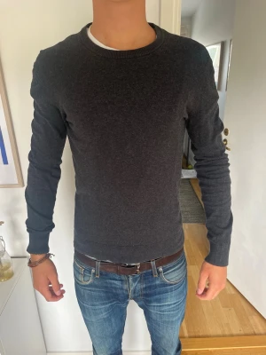 Mörkgrå rundhalsad tröja - Snygg och enkel mörkgrå tröja med rund halsringning och långa ärmar. Tröjan har en smal passform och ribbade muddar vid ärmslut och nederkant. Perfekt att matcha med jeans för en clean look. Model är 185 cm, väger 71kg. Hör av er för mer info