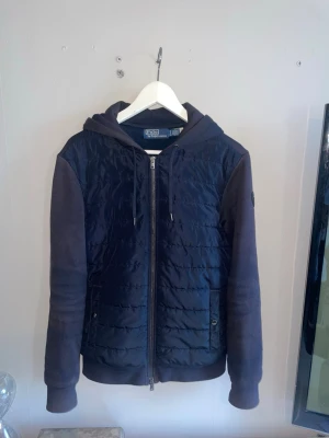 Mörkblå cardigan från Ralph Lauren - Snygg mörkblå cardiganjacka med huva och dragkedja framtill. Framsidan är quiltad medan ärmar och baksida är i mjukt sweatshirtmaterial. Två fickor med knapp framtill och ribbade muddar vid ärmslut och nederkant. Perfekt för lager på lager.