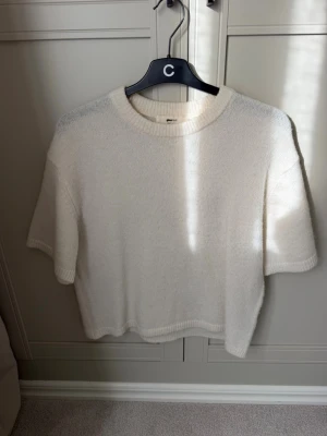 Vit stickad t-shirt med kort ärm - Säljer en enkel och stilren vit stickad t-shirt med korta ärmar och rund hals. Tröjan har en rak passform och ribbade kanter vid hals, ärmslut och nederkant. Perfekt att matcha med jeans eller kjol för en clean look.