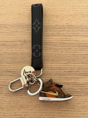  Vuitton nyckelring sneaker - Snygg nyckelring från  Vuitton med svart läderrem och klassiskt monogrammönster. Hängande detalj i form av en brun miniatyrsneaker med vita och orange detaljer samt LV-logga. Metallringar och karbinhake i silverfärg.